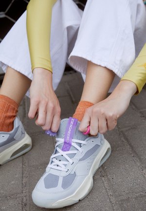 Persona con calcetines naranjas y pantalones blancos atándose zapatillas grises y blancas con lengüetas moradas al aire libre sobre una superficie pavimentada.