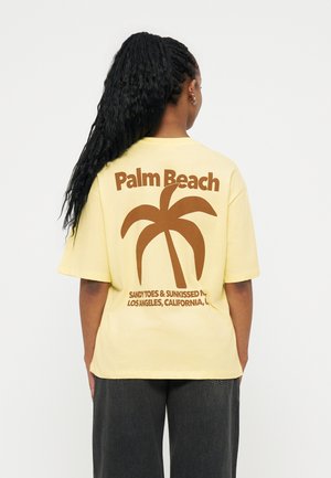 Άτομο με μακρύ πλεγμένο μαλλί, φορώντας κίτρινο T-shirt με καφέ κείμενο "Palm Beach" και γραφικό φοίνικα στην πλάτη.