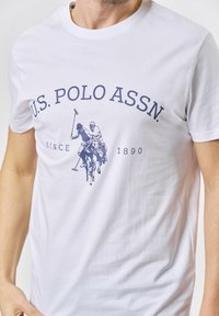 Baltas medvilnės marškinėliai su mėlynu piešiniu, kuriame pavaizduoti du polo žaidėjai, ir tekstu "U.S. POLO ASSN. SINCE 1890" tamsiai mėlynos spalvos. Apskritas kaklas.