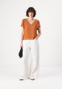 Top de color óxido de manga corta con escote en V y mangas de volantes, combinado con pantalones de pierna ancha blancos y un bolso negro. Zapatos planos con ribete de contraste.