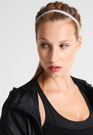 Giacca sportiva nera, canotta nera sotto, e una sottile fascia bianca. Focus sul viso con capelli intrecciati, trucco naturale e sfondo neutro.