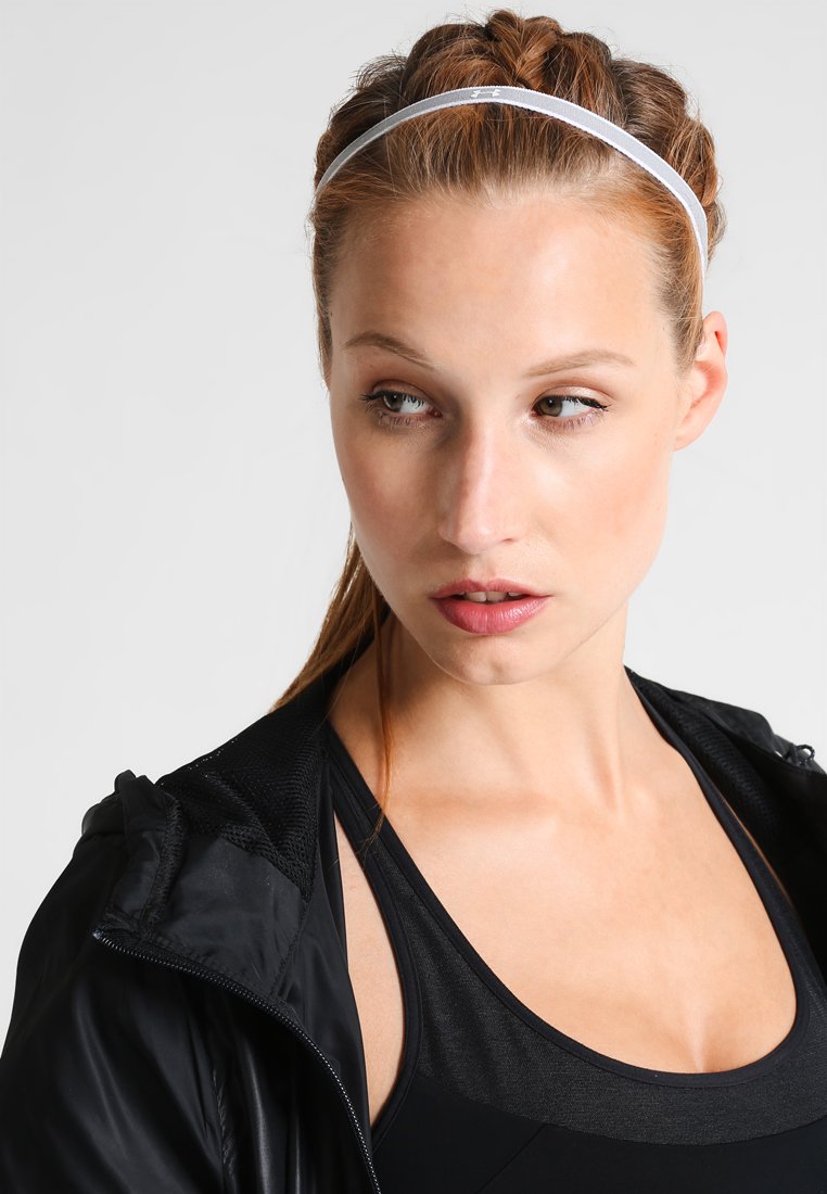 Giacca sportiva nera, canotta nera sotto, e una sottile fascia bianca. Focus sul viso con capelli intrecciati, trucco naturale e sfondo neutro.