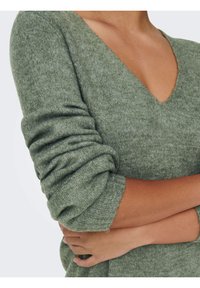 Pull à col en V à manches longues en tissu vert doux et texturé avec manches retroussées et poignets côtelés.