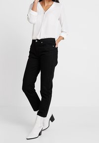 Černé tapered džíny s vysokým pasem, pětikomorový design, denimový materiál, spárované s bílou blůzkou a bílými polobotkami na podpatku.