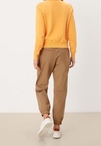 Orange strikketrøje, tan elastiske joggers med lynlåslommer og hvide sneakers, set fra ryggen mod en neutral baggrund.