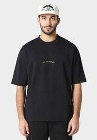 Black cotton t-shirt with short sleeves, features gold embroidered text "PAS DE MONACO" centered on the chest, relaxed fit, minimalistic design.