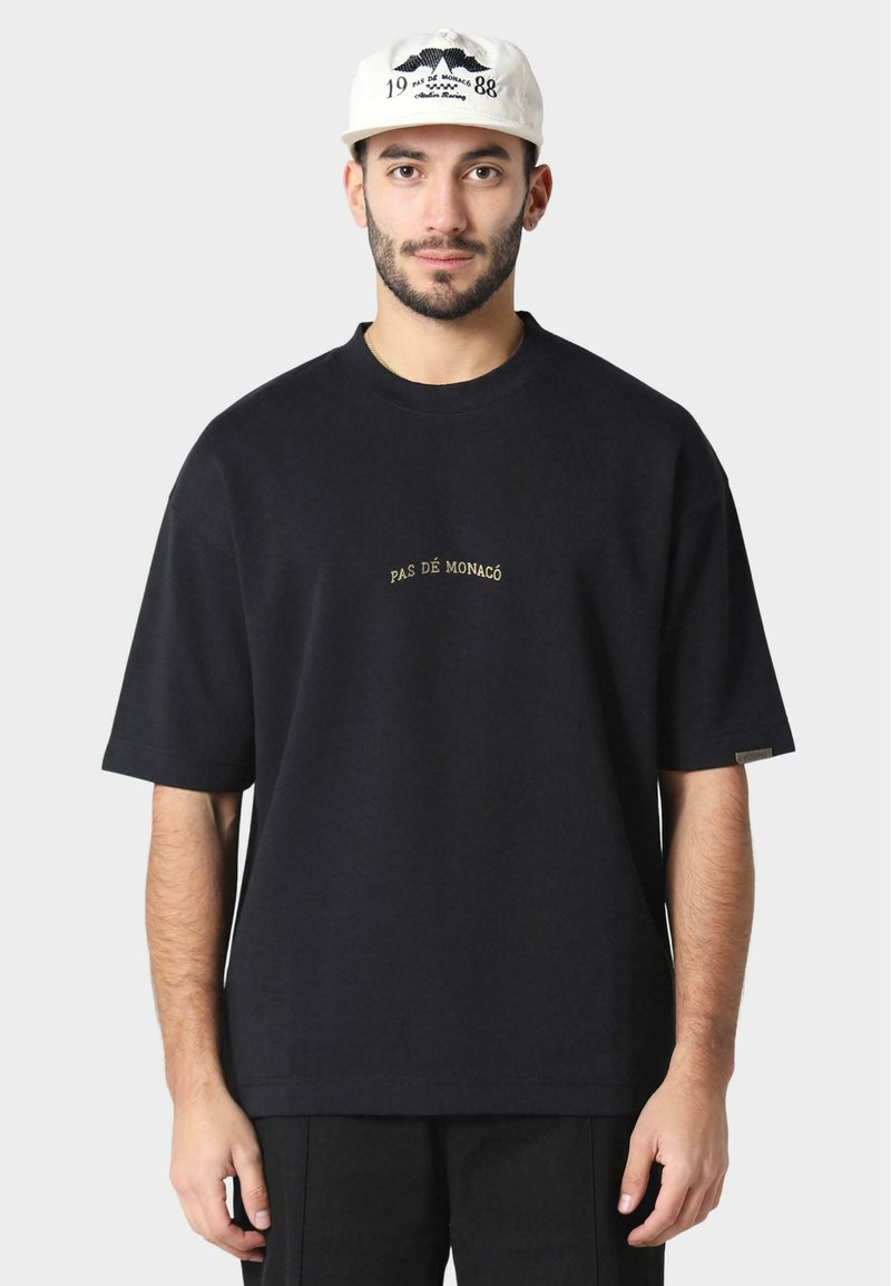 Black cotton t-shirt with short sleeves, features gold embroidered text "PAS DE MONACO" centered on the chest, relaxed fit, minimalistic design.