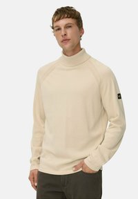 Junger Mann in einem beigen Rollkragenpullover mit langen Ärmeln und dunkler Hose, mit einer Hand in seiner Tasche vor einem schlichten Hintergrund.