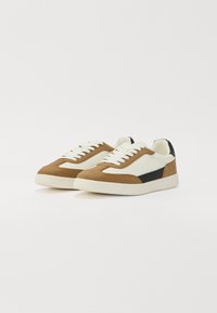 Baskets beige et blanches avec une touche de noir. Fabriquées en suede et en cuir, elles présentent un laçage à l'avant et une semelle texturée. Design classique.