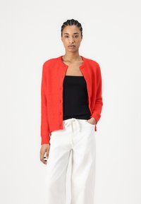 Red knitted cardigan with round neckline, button closure, ribbed cuffs, worn over a black tank top, paired with white wide-leg trousers.