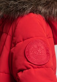 Rode pufferjas met een bontgevoerde capuchon. Bevat een gemarmerde patch met berggraphics op de linkermouw. Glad materiaal met gewatteerde delen.