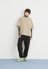 Béžové oversized tričko, černé kalhoty a zelenou obuv. Materiál vypadá měkce; tričko má jemné logo; volný střih s mírně spadlými rameny.