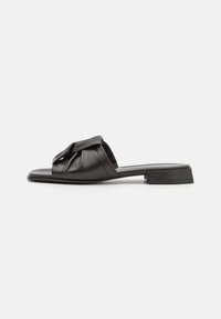 Svart lädersandal med halkfri sula som har en knuten design framtill och en låg klack för stabilitet. Slät yta och minimalistisk form.