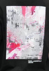 Čierne bavlnené tričko s abstraktným dizajnom v šedej a ružovej farbe, vrátane škvŕn a kvapiek. Obsahuje logo "KARL LAGERFELD JEANS."