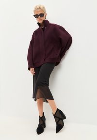 Giacca oversize bordeaux con colletto alto, gonna nera trasparente e stivaletti neri a punta con cinturini incrociati, su uno sfondo semplice.