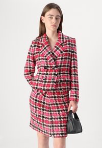 Anzug im Muster von pinkem, schwarzem und weißem Hahnentritt mit einem doppeltreihigen Blazer und einem figurbetonten Bleistiftrock; strukturierter Stoff; kombiniert mit einer schwarzen Handtasche.