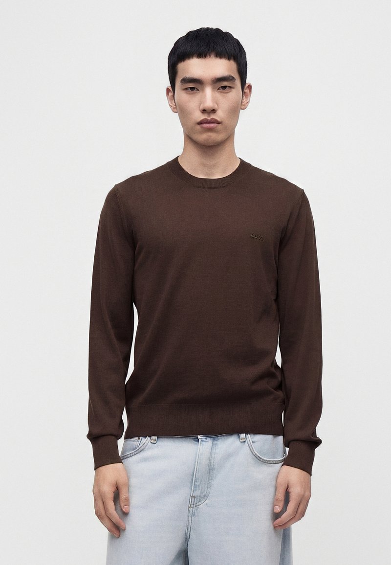 Νέος άνδρας με κοντά μαύρα μαλλιά που φοράει απλό σκούρο καφέ μακρυμάνικο πουλόβερ και ανοιχτό μπλε τζιν, στέκεται μπροστά σε απλό φωτεινό φόντο.