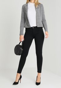 Grauer kurzer Blazer, weißes Oberteil, schwarze eng anliegende Hose, schwarze hohe Absätze und eine runde schwarze Handtasche. Glatte Texturen und strukturiertes Design.
