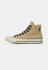 Basket haute beige avec des lacets noirs, embout en caoutchouc blanc et tige en toile texturée. Présente une bande noire le long de la semelle.