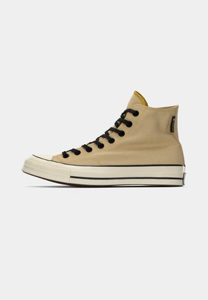 Beige hoge sneakers met zwarte veters, een witte rubberen neuskap en een gestructureerde canvas bovenkant. Heeft een zwarte streep langs de zool.