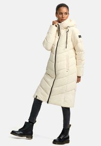 Long, cream puffer coat with a hood, featuring chevron stitching, black zipper, and two side pockets. Paired with black lace-up boots.
