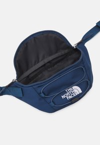 Borsa in vita di stoffa blu con interno nero, cordino elastico anteriore e logo bianco "THE NORTH FACE" sulla tasca frontale.