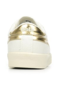 Sneaker bianco in tela con una striscia metallica dorata e un accento sul tallone. Presenta una parte superiore testurizzata e una punta arrotondata. Suola in gomma.
