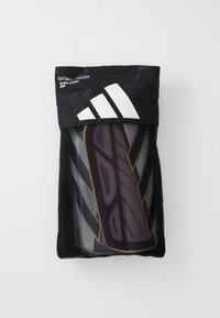 Black mesh bag containing purple and gold football shin guards. Bag features a large white Adidas logo at the top.