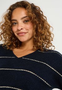 Pull bleu marine avec des rayures crémeuses en diagonale, col en V, tricot texturé et manches amples. Tissu doux et décontracté.