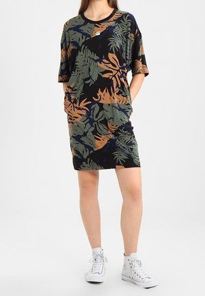 Femme portant une robe courte à manches courtes, longueur genou, avec des motifs de feuilles et abstraits en vert, marron et bleu marine, associée à des baskets blanches montantes.