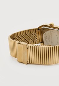 Ρολόι χειρός από μέταλλο με χρυσό τόνο, με βραχιόλι από πλέγμα και κούμπωμα, εμφανίζοντας την πίσω πλευρά χαραγμένη με την επωνυμία «Lacoste» και λεπτομέρειες μοντέλου.