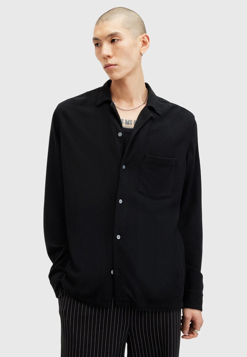 Chemise noire à boutons, en tissu doux, avec col, poche poitrine unique et manches longues. Le modèle porte un pantalon rayé.