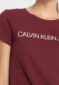 Camiseta de algodón color burdeos con cuello redondo y mangas cortas, con "CALVIN KLEIN" impreso en blanco en el pecho.