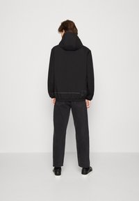 Svart hooded jacka med elastisk nederkant, slät textur och subtilt reflekterande linjedetalj. Bärs med mörka jeans och svarta sneakers.