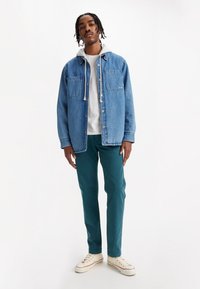 Jachetă din denim de culoare albastră deschisă, cu două buzunare pe piept, suprapusă peste un hanorac gri. Pantaloni verzi și adidași bej completează ținuta.