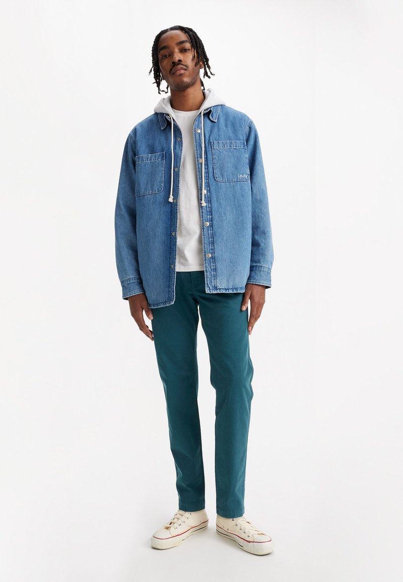 Veste en denim bleu clair avec deux poches poitrine, superposée à un hoodie gris. Un pantalon vert et des baskets beige complètent la tenue.