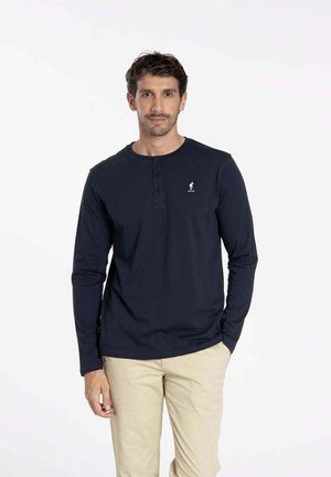 Chemise henley à manches longues bleu marine avec un col ras du cou, trois boutons au niveau du col et un petit logo sur la poitrine. En matériau coton, design ajusté.