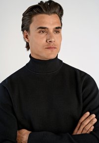 Schwarzer Rollkragensweater mit glatter Textur, tailliertem Schnitt und geripptem Kragen. Das Modell posiert mit verschränkten Armen vor einem hellen Hintergrund.