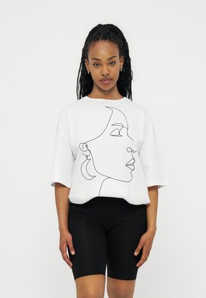 Νέα γυναίκα με μακριά πλεγμένα μαλλιά, φορώντας μια λευκή υπερμεγέθη μπλούζα με μινιμαλιστικό σχέδιο γραμμής προσώπου και μαύρα ποδηλατικά σορτς.