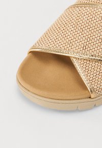 Beige kudottu sandaali, jossa on ristikkäinen muotoilu ja kultaiset yksityiskohdat sekä pehmeä, tyynyttämätön pohja. Kumipohja, jossa on tekstuuripintaisia yksityiskohtia.