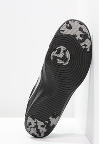 Suola di scarpa sportiva nera con un motivo testurizzato e rigato. Presenta un accento mimetico in grigio e nero. Elemento di design circolare vicino al centro.