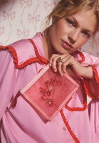 Růžová taštička s červenou výšivkou a zipem, zdobená květinovými a srdíčkovými motivy, držená před růžovou blůzou s červenými volánky.