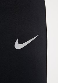 Zwarte sportlegging van rekbare stof, met een opvallend grijs Nike-logo aan de onderkant links. Gladde textuur, aansluitend ontwerp.