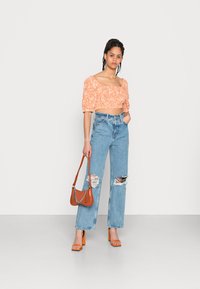 Top cropped floral laranja com mangas bufantes, jeans de perna larga desfiados em azul claro, sandálias laranjas e uma bolsa castanha com detalhes em corrente.