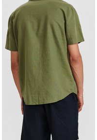 Persona che indossa una camicia verde oliva a maniche corte e pantaloni corti neri, vista di spalle, in piedi contro uno sfondo chiaro e uniforme.