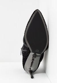 Chaussure à talon haut noire à bout pointu avec un talon aigu brillant reposant sur le côté contre un mur d'angle blanc.