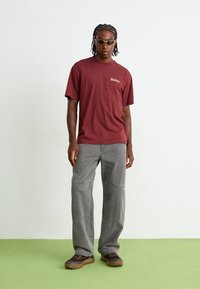 T-shirt graphique bordeaux avec le logo "Dickies", associé à un pantalon cargo gris ample et des chaussures de randonnée marron. L'homme porte des lunettes de soleil teintées vertes.
