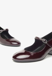 Scarpe Mary Jane in pelle verniciata color bordeaux con punta arrotondata e tacco medio. Presentano una fascia decorativa con un accento di pietra preziosa.