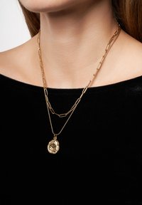 Des colliers en or laminé avec une chaîne plate et allongée et une chaîne plus courte avec un pendentif texturé. Portés sur un fond en tissu sombre.