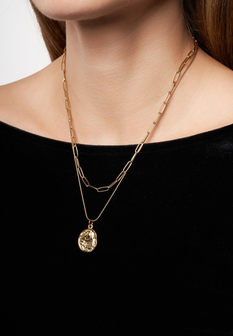 Des colliers en or laminé avec une chaîne plate et allongée et une chaîne plus courte avec un pendentif texturé. Portés sur un fond en tissu sombre.